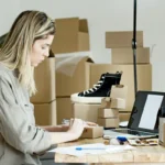 Perempuan mengemas produk untuk toko online dengan laptop di dekatnya, menggambarkan bisnis dropship dari rumah